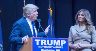 Man in a dark suit speaks at a podium with a blue 'TRUMP' campaign sign; a woman in a beige outfit stands nearby on a stage with flags in the background.