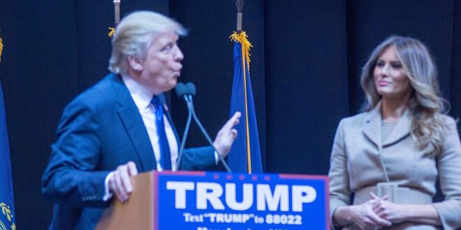 Man in a dark suit speaks at a podium with a blue 'TRUMP' campaign sign; a woman in a beige outfit stands nearby on a stage with flags in the background.