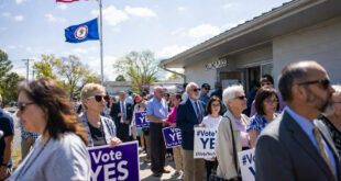 Crowd of people holding 'Vote YES' signs outside a polling place, with American and state flags waving in the sky.