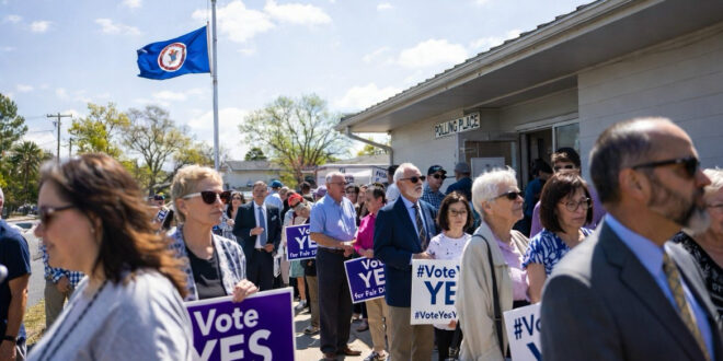 Crowd of people holding 'Vote YES' signs outside a polling place, with American and state flags waving in the sky.