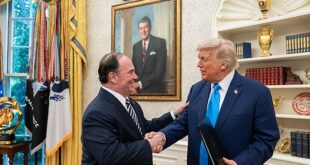 Two men in suits shake hands in a formal, gold-decorated room with flags and a framed portrait on the wall behind them.