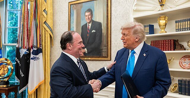 Two men in suits shake hands in a formal, gold-decorated room with flags and a framed portrait on the wall behind them.