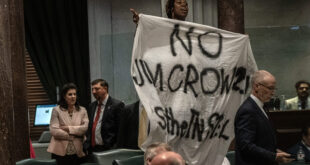 Person lifts a large white banner with dark handwritten text in a formal meeting room as suited attendees look on.