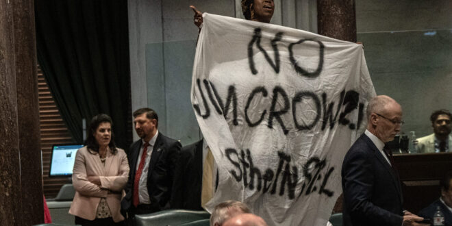 Person lifts a large white banner with dark handwritten text in a formal meeting room as suited attendees look on.