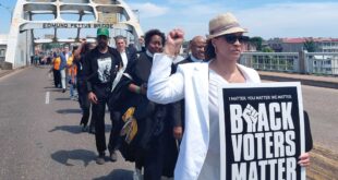 Protesters march across Edmund Pettus Bridge on a sunny day; foreground woman in white blazer and hat holds a large Black Voters Matter sign (reading 'BLACK VOTERS MATTER').