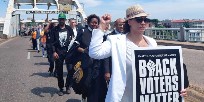Protesters march across Edmund Pettus Bridge on a sunny day; foreground woman in white blazer and hat holds a large Black Voters Matter sign (reading 'BLACK VOTERS MATTER').