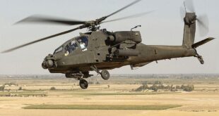 Military attack helicopter in flight over a barren desert landscape with rotors spinning.