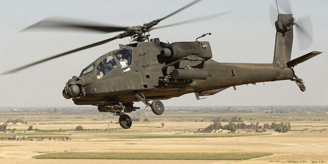 Military attack helicopter in flight over a barren desert landscape with rotors spinning.