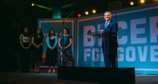 A man in a suit speaks at a clear podium on a stage, with five women standing to his left and a large banner behind him reading 'FOR GOV'.