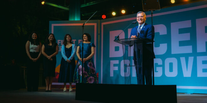 A man in a suit speaks at a clear podium on a stage, with five women standing to his left and a large banner behind him reading 'FOR GOV'.