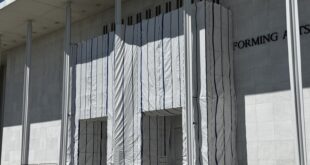 Facade of a modern building with a white fabric-covered entrance wrapped with blue vertical stripes during construction, with barricades in front