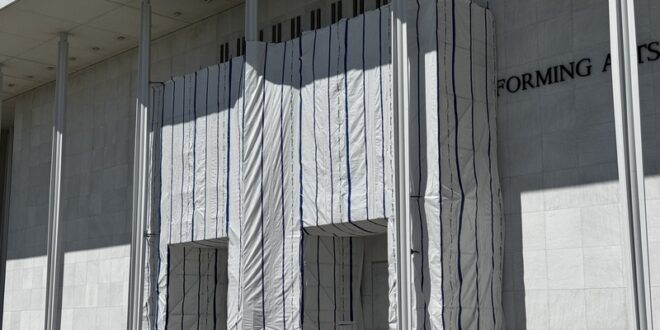 Facade of a modern building with a white fabric-covered entrance wrapped with blue vertical stripes during construction, with barricades in front