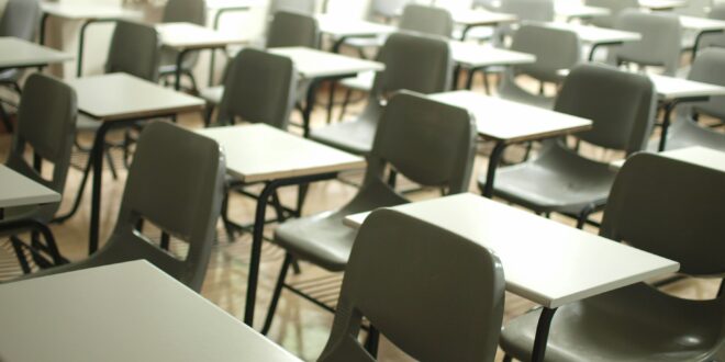Empty classroom with rows of gray desks and chairs, sunlight by the windows at the back.