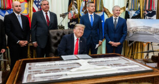 President Donald Trump sits at a large ornate desk signing a document while four suited aides stand behind him, with flags and draped curtains in the background.