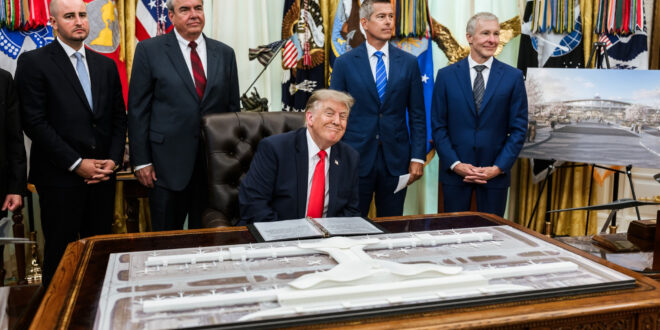 President Donald Trump sits at a large ornate desk signing a document while four suited aides stand behind him, with flags and draped curtains in the background.