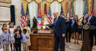 President Trump smiles beside a ceremonial bell as children applaud in a gilded room with flags in the background.