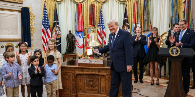 President Trump smiles beside a ceremonial bell as children applaud in a gilded room with flags in the background.