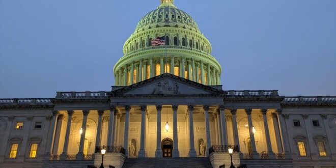 Front view of the U.S. Capitol at dusk, dome lit with a flag visible, grand staircase and columns in the foreground.