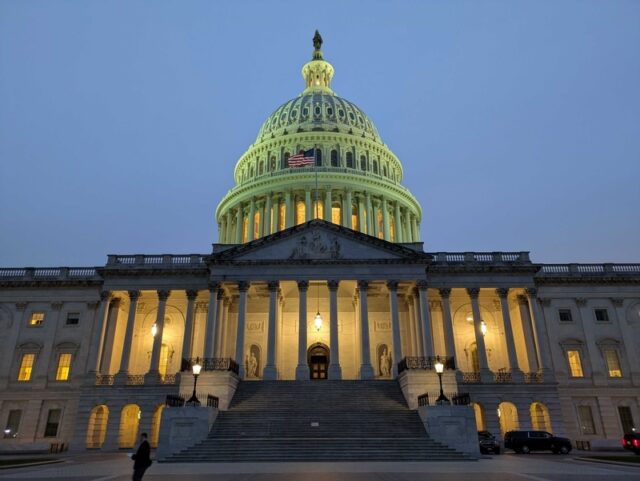 capitol at night - Poli Alert Front view of the U.S. Capitol at dusk, dome lit with a flag visible, grand staircase and columns in the foreground.