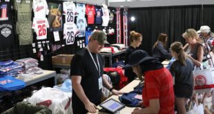 Vendor at a merchandise booth helping a customer at a registration/tablet checkout in a large tent, with shelves of t-shirts on display in the background.