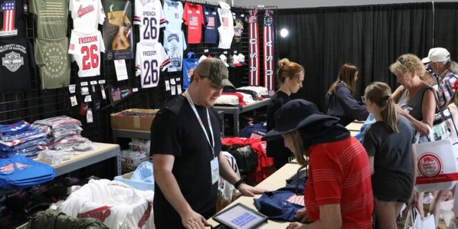 Vendor at a merchandise booth helping a customer at a registration/tablet checkout in a large tent, with shelves of t-shirts on display in the background.