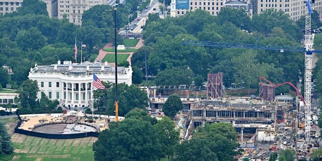 Aerial view of the White House grounds with a construction site and tall crane to the right, surrounded by trees and city buildings.