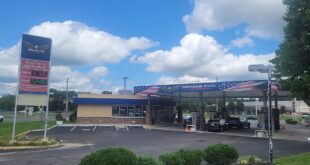 Freedom Fuel gas station with a blue, flag-themed canopy and cars fueling under a sunny, partly cloudy sky.