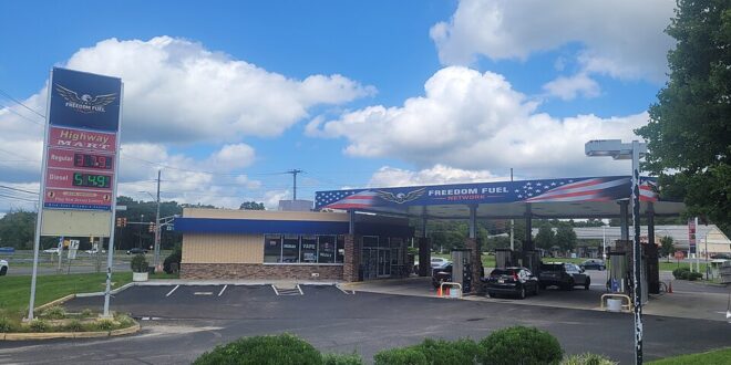 Freedom Fuel gas station with a blue, flag-themed canopy and cars fueling under a sunny, partly cloudy sky.