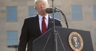 Man in a dark suit and red tie delivers a speech at a lectern with a microphone, outdoors, with the presidential seal visible on the podium.