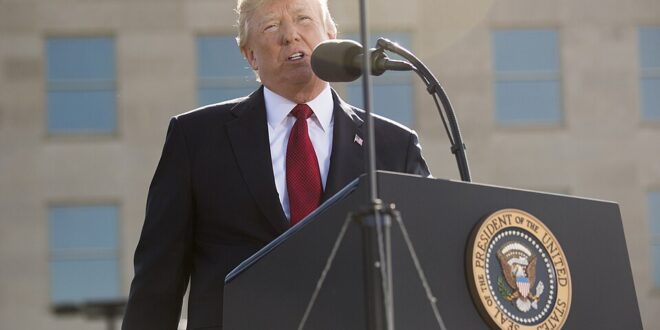 Man in a dark suit and red tie delivers a speech at a lectern with a microphone, outdoors, with the presidential seal visible on the podium.