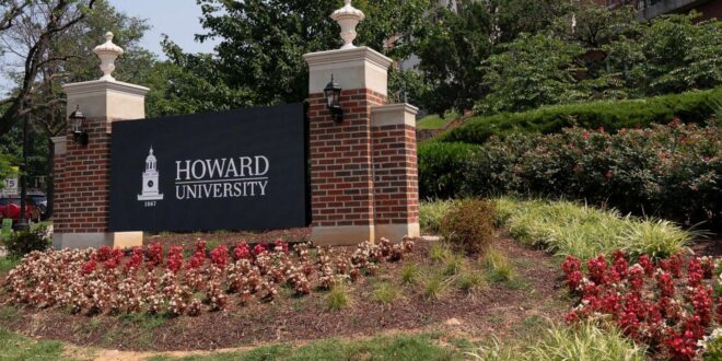 Howard University entrance sign flanked by brick pillars, lanterns, and blooming flower beds in a landscaped campus entrance.