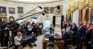President Trump seated at a large desk in a gold-edged, ornate room, with reporters, photographers, and microphones overhead as officials stand nearby.