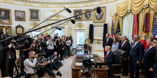 President Trump seated at a large desk in a gold-edged, ornate room, with reporters, photographers, and microphones overhead as officials stand nearby.