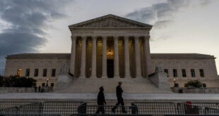 U.S. Supreme Court Building at dusk with tall columns and steps, two people walking past security barriers in the foreground.