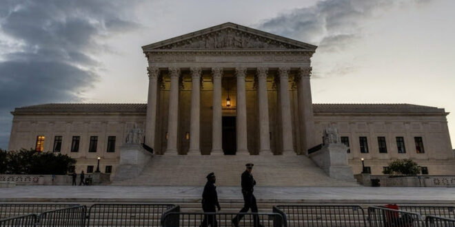 U.S. Supreme Court Building at dusk with tall columns and steps, two people walking past security barriers in the foreground.