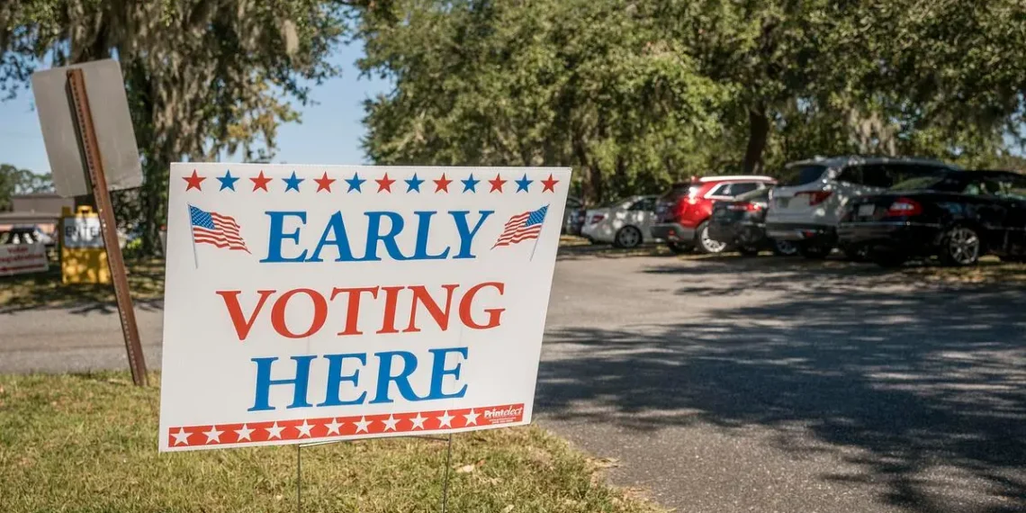 20241017-early-voting-107jpg - Poli Alert Election sign reading 'Early Voting Here' placed on a grassy verge beside a parking lot with cars and trees in the background.