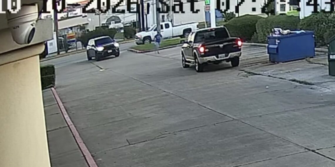 D58F597B-9C29-4358-BCC3-81972CC49948 - Poli Alert Surveillance camera view of a parking lot with a black pickup in the center, a person walking in the background, and a blue dumpster on the right.