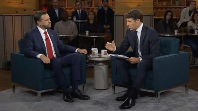 Two men in dark suits sit in blue armchairs on a talk-show set, host on the right with papers, guest on the left; audience in background